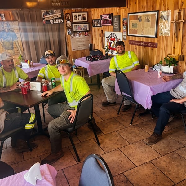 Six men in work attire are seated around tables with purple tablecloths in a casual restaurant.