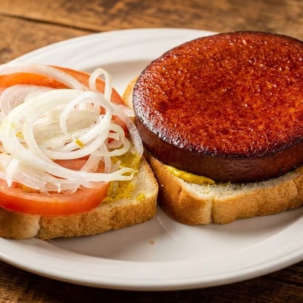 A fried bologna sandwich with mustard, tomato, and onion on white bread, on a white plate.