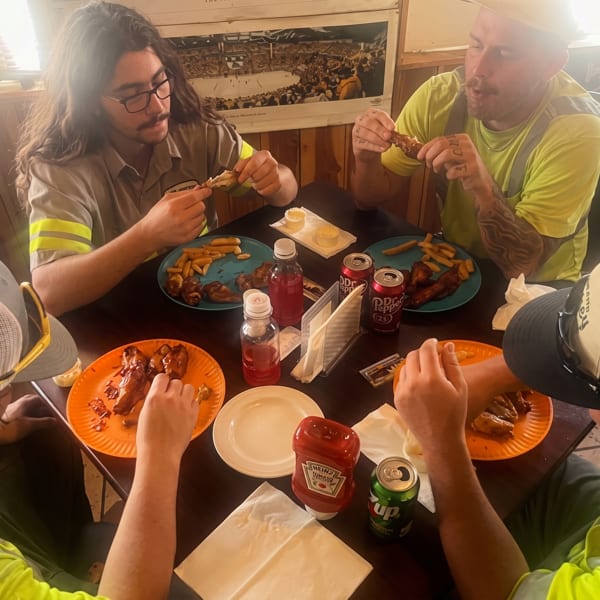Four men in work attire eating chicken wings and fries at a dark wooden table.