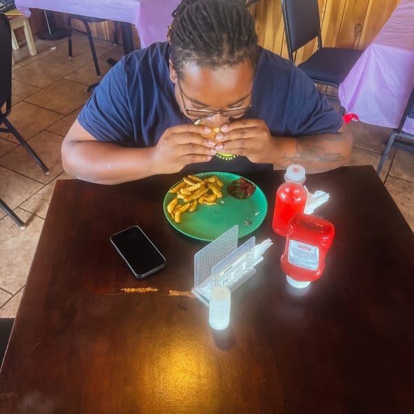A person with dreadlocks eating a burger and fries at a wooden table in a casual restaurant.