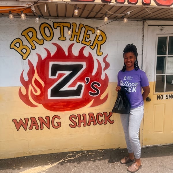 A smiling woman stands in front of Brother Z's Wang Shack, holding a black bag.