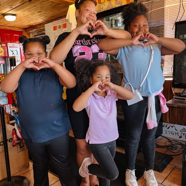 A woman and three girls pose making heart shapes with their hands inside a restaurant.